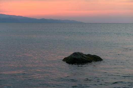 A serene view of calm waters reflecting the soft hues of sunset, with a solitary rock and distant mountains.