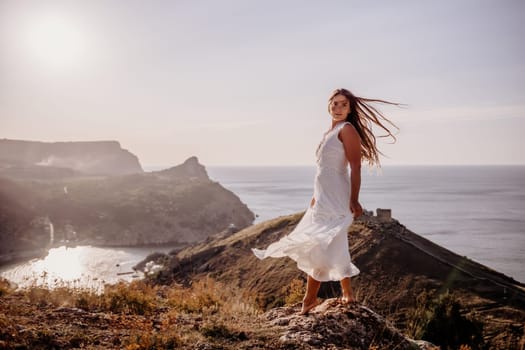 A woman in a white dress stands on a hill overlooking the ocean. The scene is serene and peaceful, with the woman's dress billowing in the wind. The combination of the ocean