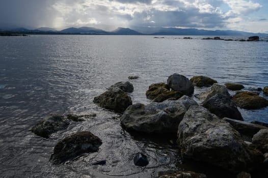 Gentle waves lap against rugged rocks while dark clouds loom above a peaceful waterway at dusk.