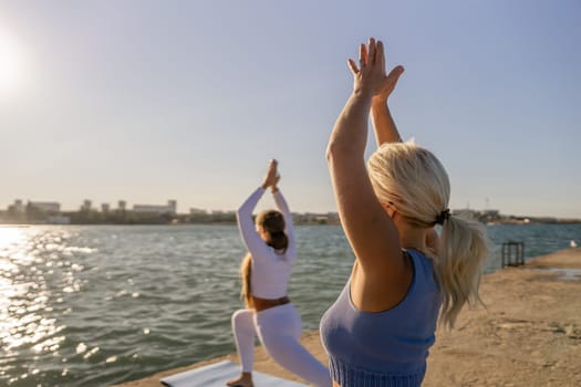 Two women practice yoga poses on a pier overlooking a calm body of water. The woman in the foreground is blonde and wearing a blue tank top.