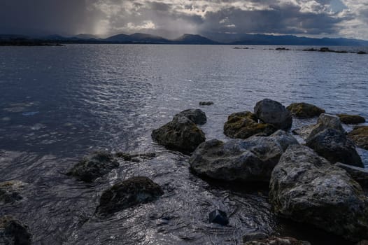 Dark clouds loom over the tranquil lake, casting shadows on the rocky shoreline as the sun begins to set.