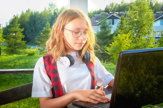 Girl with headphones focuses on laptop screen in a serene outdoor setting, surrounded by greenery on a sunny day.