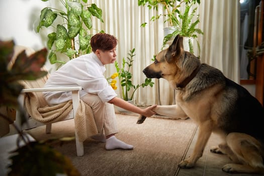 A woman sits with her German Shepherd in a comfortable space filled with plants, enjoying a quiet moment together.