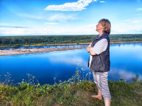 A middle-aged blonde woman takes a selfie while standing barefoot by the river, appreciating the beautiful landscape.