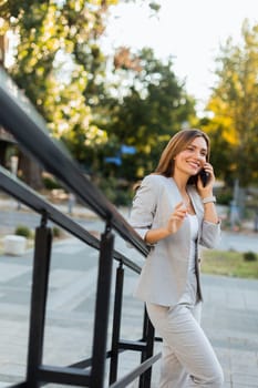 Woman strolls outdoors in a chic gray suit, animatedly chatting on her phone. The natural light enhances her radiant smile, surrounded by beautiful greenery