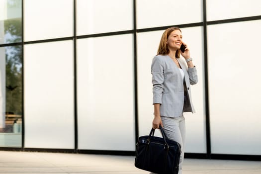 Professional woman strolls confidently along a sleek glass structure, her expression joyful as she chats on the phone, dressed in a stylish gray business suit