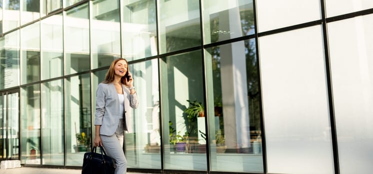 Professional woman strolls confidently along a sleek glass structure, her expression joyful as she chats on the phone, dressed in a stylish gray business suit