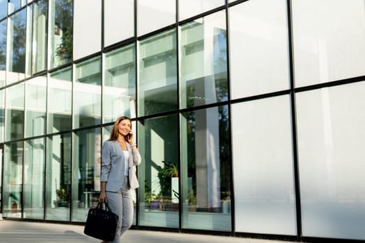 Professional woman strolls confidently along a sleek glass structure, her expression joyful as she chats on the phone, dressed in a stylish gray business suit