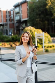 Confident young woman in a light gray suit engages with her smartphone, radiating positivity as she enjoys the lively urban atmosphere filled with greenery