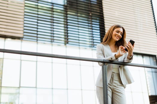 Cheerful business woman stands on a balcony, engaged with her smartphone against a backdrop of modern architecture, soaking in the warm sunlight and vibrant atmosphere