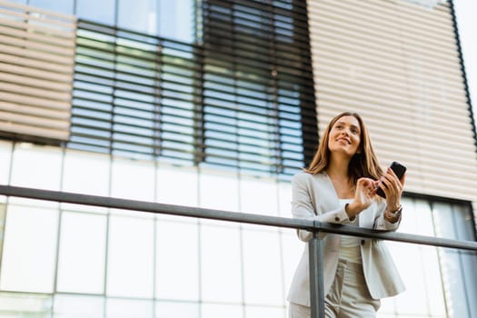 Cheerful business woman stands on a balcony, engaged with her smartphone against a backdrop of modern architecture, soaking in the warm sunlight and vibrant atmosphere