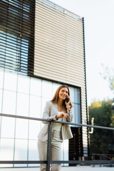 Cheerful business woman stands on a balcony, engaged with her smartphone against a backdrop of modern architecture, soaking in the warm sunlight and vibrant atmosphere