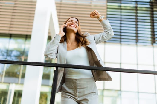 Joyful business woman celebrates a moment outdoors, engaging in a cheerful phone conversation while leaning against a railing beside an elegant building.