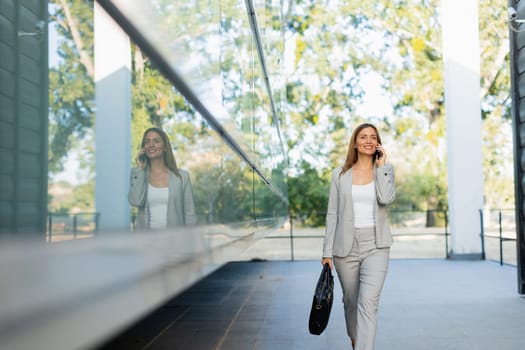 With a warm smile, a professional woman walks confidently along a sleek corridor, chatting on her phone as the sunlight filters through the trees