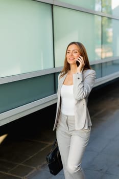 With a warm smile, a professional woman walks confidently along a sleek corridor, chatting on her phone as the sunlight filters through the trees