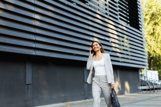 Walking with purpose, a business woman in a stylish outfit enjoys a phone conversation on a bright day, showcasing her confident demeanor against a sleek architectural backdrop