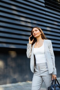 Walking with purpose, a business woman in a stylish outfit enjoys a phone conversation on a bright day, showcasing her confident demeanor against a sleek architectural backdrop