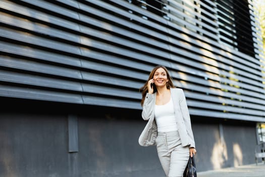 Walking with purpose, a business woman in a stylish outfit enjoys a phone conversation on a bright day, showcasing her confident demeanor against a sleek architectural backdrop