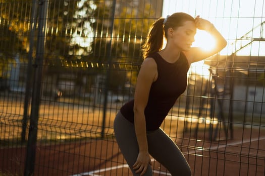 In the golden glow of sunset, a focused woman leans against a fence, catching her breath after an energizing workout, embodying strength and persistence