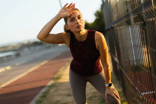 Woman pauses during her evening workout, wiping her brow as the sun sets, surrounded by a tranquil jogging path and a softly lit landscape