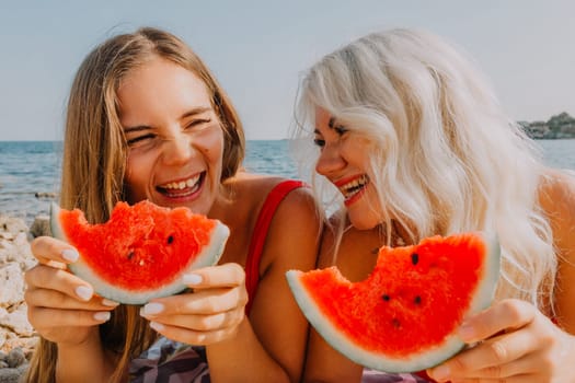 Two women, one young with long blonde hair, the other older with white hair, joyfully eat watermelon slices on a beach. Sunlight brightens their happy faces.