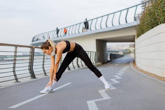 A fit woman wearing athletic clothing performs a stretching routine on a riverside pathway, with serene water views and leisurely walkers enjoying a peaceful morning outside.