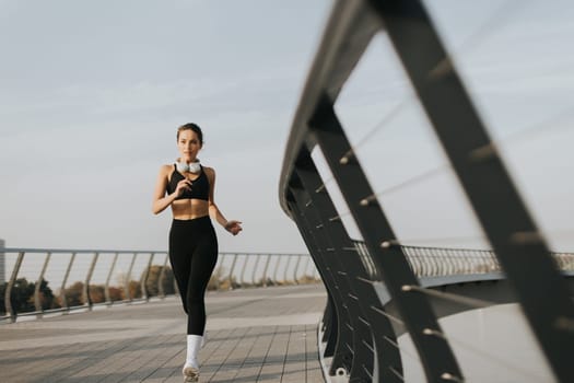 Fit woman in a black athletic outfit runs on a sleek bridge, enjoying her evening routine. The horizon glows softly, highlighting her determination and commitment to fitness