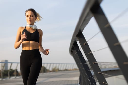 Fit woman in a black athletic outfit runs on a sleek bridge, enjoying her evening routine. The horizon glows softly, highlighting her determination and commitment to fitness