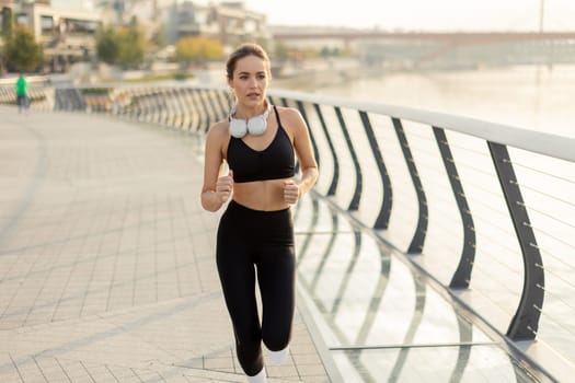 Fit woman in a black athletic outfit runs on a sleek bridge, enjoying her evening routine. The horizon glows softly, highlighting her determination and commitment to fitness