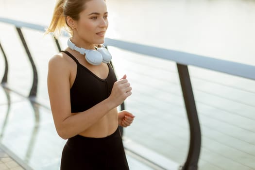 A young woman in athletic attire runs energetically along a riverside path, embracing the serene atmosphere of a beautiful morning, fueled by her favorite music.