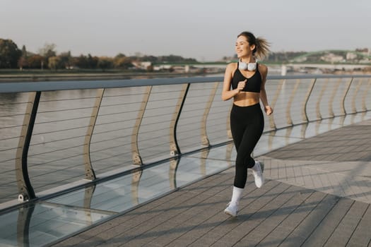 A young woman in athletic attire runs energetically along a riverside path, embracing the serene atmosphere of a beautiful morning, fueled by her favorite music.