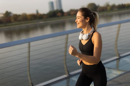 A young woman in athletic attire runs energetically along a riverside path, embracing the serene atmosphere of a beautiful morning, fueled by her favorite music.