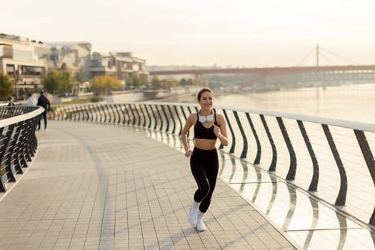 A young woman in athletic attire runs energetically along a riverside path, embracing the serene atmosphere of a beautiful morning, fueled by her favorite music.
