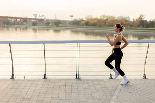 A determined runner strides confidently along a riverside path, embracing the crisp morning air, with city skyline and autumn trees providing a picturesque backdrop.