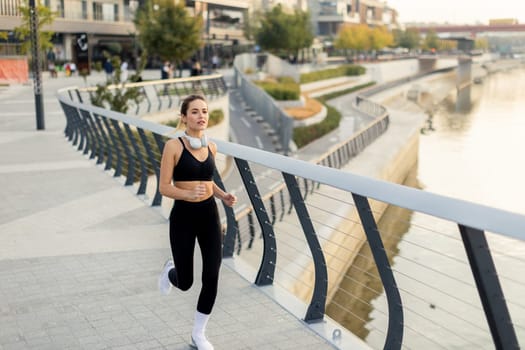 A dedicated runner maintains her pace on a riverwalk surrounded by urban architecture and lush greenery, embracing a beautiful morning and an active lifestyle.