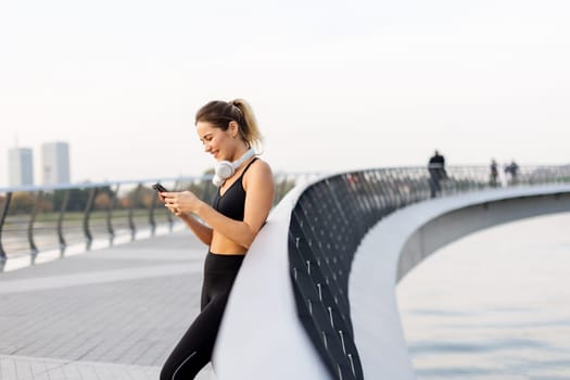 A jogger pauses to chat on her phone by the riverside, wearing workout gear and headphones, embracing the warm sunshine and the serene atmosphere around her.
