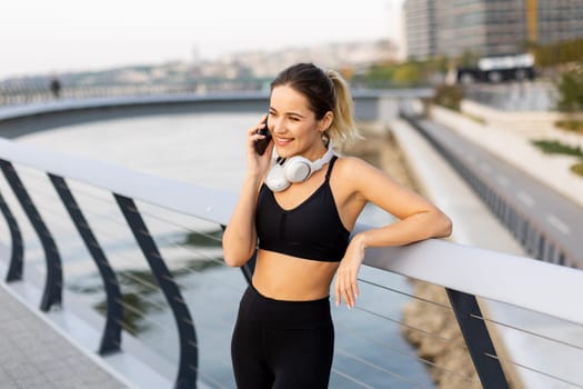 A jogger pauses to chat on her phone by the riverside, wearing workout gear and headphones, embracing the warm sunshine and the serene atmosphere around her.