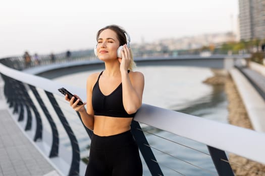 A young woman stands relaxed on a bridge, headphones on, savoring music while basking in the gentle glow of sunset near the water's edge.