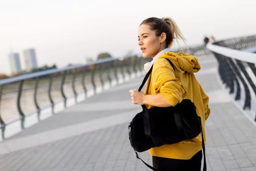 Walking along the riverside path, a cheerful woman in a yellow hoodie beams with joy. The evening sky casts a warm glow, creating a serene atmosphere for a leisurely stroll
