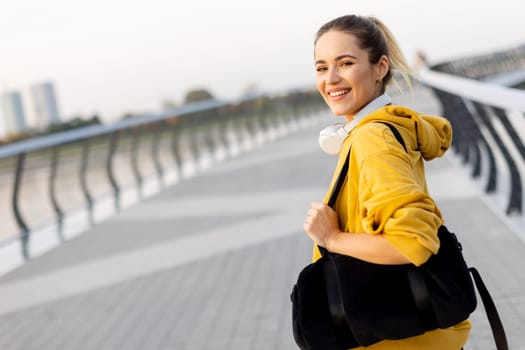 With a joyful smile, a young woman in a yellow hoodie strolls along a scenic riverside path, her black bag slung over her shoulder, embracing the gentle evening breeze.