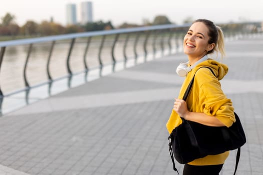 A cheerful woman strolls along a picturesque waterfront pathway, wearing a bright yellow hoodie and headphones. Her vibrant smile reflects the sunny ambiance of the day.
