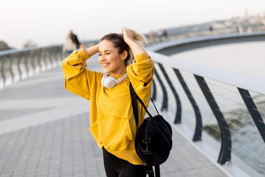 A woman in a yellow sweatshirt smiles brightly as she adjusts her ponytail, enjoying the warm sunlight by the water’s edge, immersed in her favorite tunes.
