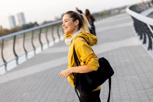 Walking along the riverside path, a cheerful woman in a yellow hoodie beams with joy. The evening sky casts a warm glow, creating a serene atmosphere for a leisurely stroll
