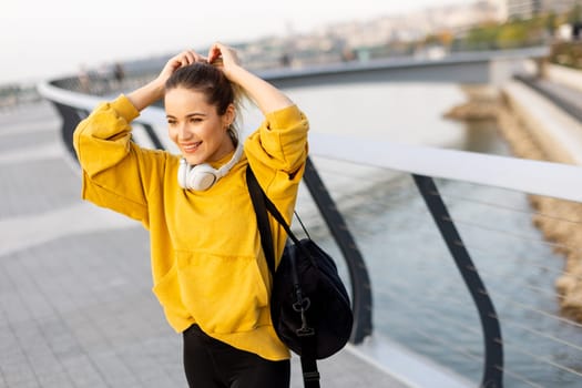 Walking along the riverside path, a cheerful woman in a yellow hoodie beams with joy. The evening sky casts a warm glow, creating a serene atmosphere for a leisurely stroll