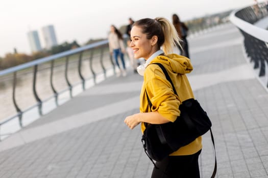 Walking along the riverside path, a cheerful woman in a yellow hoodie beams with joy. The evening sky casts a warm glow, creating a serene atmosphere for a leisurely stroll