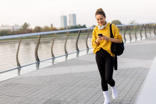 As the sun rises over the skyline, a young woman dressed in a vibrant yellow sweater walks alongside the river, captivated by her smartphone while listening to music.