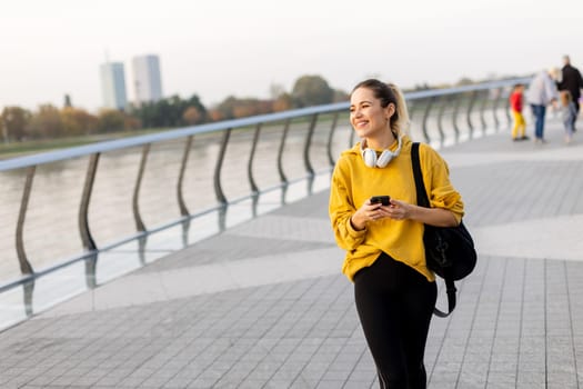 A smiling young woman walks along a riverside path in autumn, dressed in a cozy yellow sweater and headphones. The serene atmosphere captures her joyful moment.