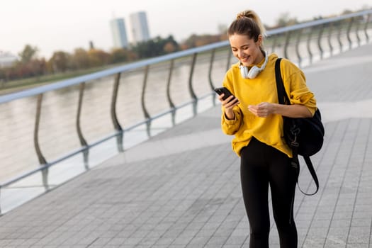 Joyful young woman strolls along a riverside walkway, engaged in a lively phone conversation while wearing a bright yellow sweatshirt and carrying a black bag