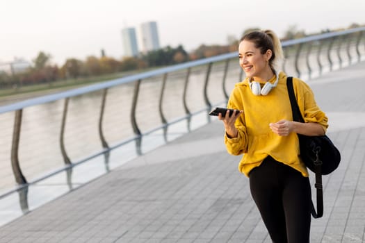Joyful young woman strolls along a riverside walkway, engaged in a lively phone conversation while wearing a bright yellow sweatshirt and carrying a black bag