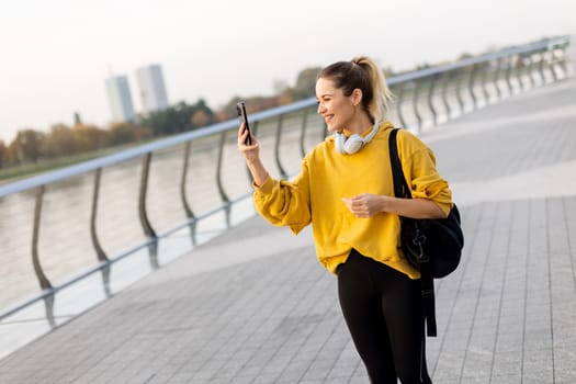 Joyful young woman strolls along a riverside walkway, engaged in a lively phone conversation while wearing a bright yellow sweatshirt and carrying a black bag